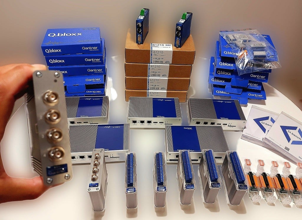 Sortierte Gantner Instruments Q.bloxx und verwandte elektronische Messmodule auf einem Tisch, einschließlich Schnittstelleneinheiten, verpackte Komponenten und Steckverbinder